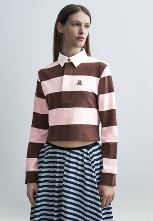 Žena s dlhými rovnými vlasmi, oblečená v hnedom a ružovom pruhovanom tričku s dlhým rukávom a bielym golierom, a v modro-bielo pruhovanej sukni.