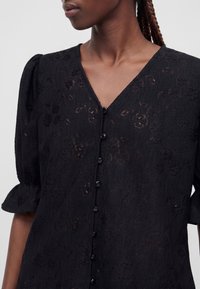 Femme portant un chemisier noir texturé à motifs floraux, boutonné devant, manches bouffantes et encolure en V, légèrement tournée vers la gauche.