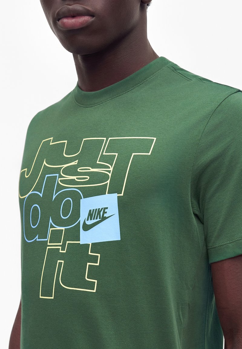 Mand iført en grøn Nike T-shirt med teksten "Just do it" i gul og blå samt en lille blå firkant med Nike-logo på brystet.