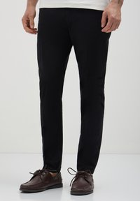 Homme portant un pantalon noir ajusté et des mocassins en cuir marron à lacets, se tenant devant un fond clair et uni.