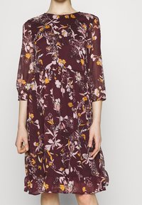 Viininpunainen kukkakuvioinen mekko, jossa on kolmen neljänneksen hihat, vaaleanpunaisia ja oransseja kukkia, röyhelöhelma ja kevyt, laskeutuva kangas.