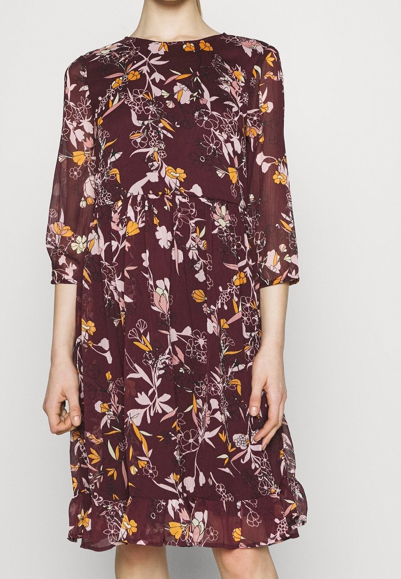 Viininpunainen kukkakuvioinen mekko, jossa on kolmen neljänneksen hihat, vaaleanpunaisia ja oransseja kukkia, röyhelöhelma ja kevyt, laskeutuva kangas.