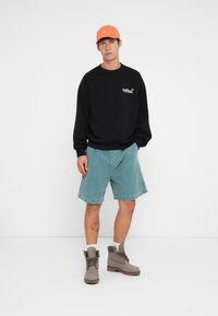 Zwarte sweatshirt met logo, lichtblauwe denim shorts, grijze laarzen en een oranje pet. Model staat tegen een witte achtergrond.