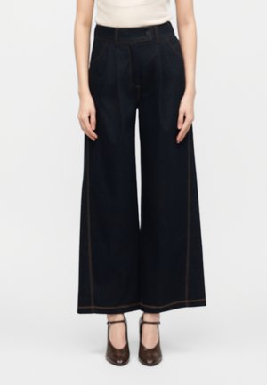 Kvinde iført sort bukser med høj talje og vide ben med brune syninger, en lys ærmeløs top og mørkebrune sko med ankelrem.