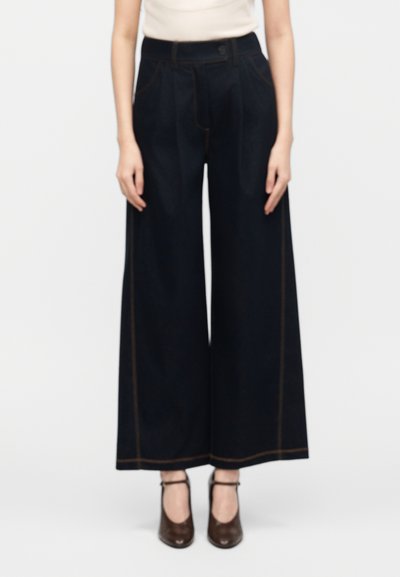 Femme portant un pantalon noir taille haute à jambes larges avec des coutures marron, un haut léger sans manches, et des chaussures à bride à la cheville marron foncé.