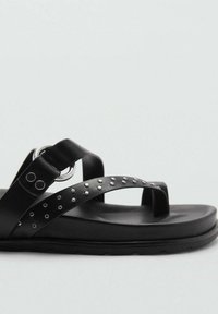 Černé ploché sandály s dvěma řemeny z kůže se hřebíky, kovovým kroužkem a polstrovanou podrážkou na jednoduchém světlém pozadí.