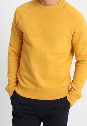 Mand iført en sennepsgul langærmet sweatshirt med hænderne i lommerne og mørkeblå bukser.