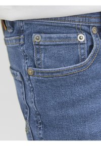 Jean en denim bleu avec des éléments en métal argenté, comportant une poche avant avec un bouton décoratif. Le tissu a une finition en denim classique et texturée.