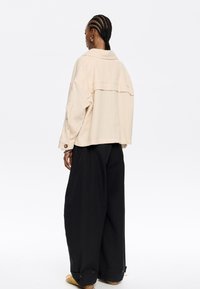 Giacca oversized beige con dettaglio della cucitura sul retro, abbinata a pantaloni neri larghi. La giacca presenta un colletto e maniche con bottoni.