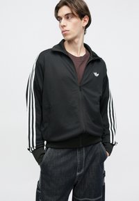 Μαύρο τζάκετ με φερμουάρ, κατασκευασμένο από υφασμά με υφή, με τρεις λευκές ρίγες σε κάθε μανίκι και μικρό λευκό λογότυπο στο αριστερό στήθος.