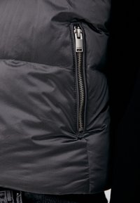 Giacca imbottita grigia con motivo trapuntato diagonale, dotata di tasca con cerniera metallica sul lato sinistro e texture liscia e lucida.