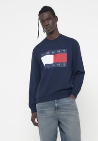 Felpa blu navy con un grande logo grafico rosso, bianco e blu. Presenta un colletto a girocollo e polsini a coste. Indossata con jeans blu chiaro.