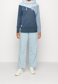 Felpa color block blu e azzurro con un motivo a zigzag sul cappuccio, abbinata a jeans larghi in denim chiaro e sneakers bianche.