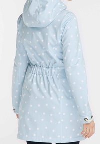 Femme portant un imperméable à capuche bleu clair à pois blancs, taille élastique et détails avec cordons aux poignets.