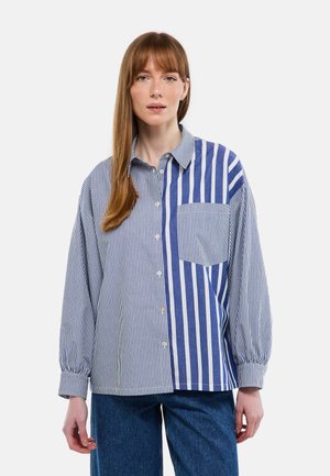 Femme portant une chemise à rayures verticales bleues et blanches avec une poche avant, associée à un jean bleu, debout devant un fond clair.