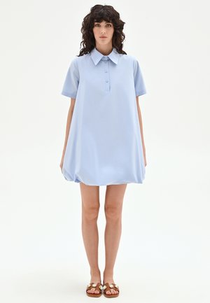 Femme aux cheveux bouclés et foncés portant une robe boutonnée à manches courtes bleu clair et des sandales marron avec des détails décoratifs, debout devant un fond blanc.