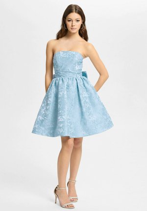 Młoda kobieta stoi ubrana w bezramkową jasnoniebieską sukienkę z kwiatowym żakardem, z rozkloszowanym dołem i wysokimi obcasami z paskiem wokół kostki, na białym tle.