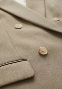 Beige skräddarsydd kavaj i en texturerad tyg, med två stora krämfärgade knappar och en notch-krage. Inkluderar eleganta fickdetaljer.