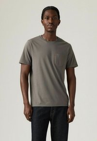 En ung mand står midt i billedet iført en grå kortærmet t-shirt med en brystlomme, parret med mørke jeans og en neutral baggrund.