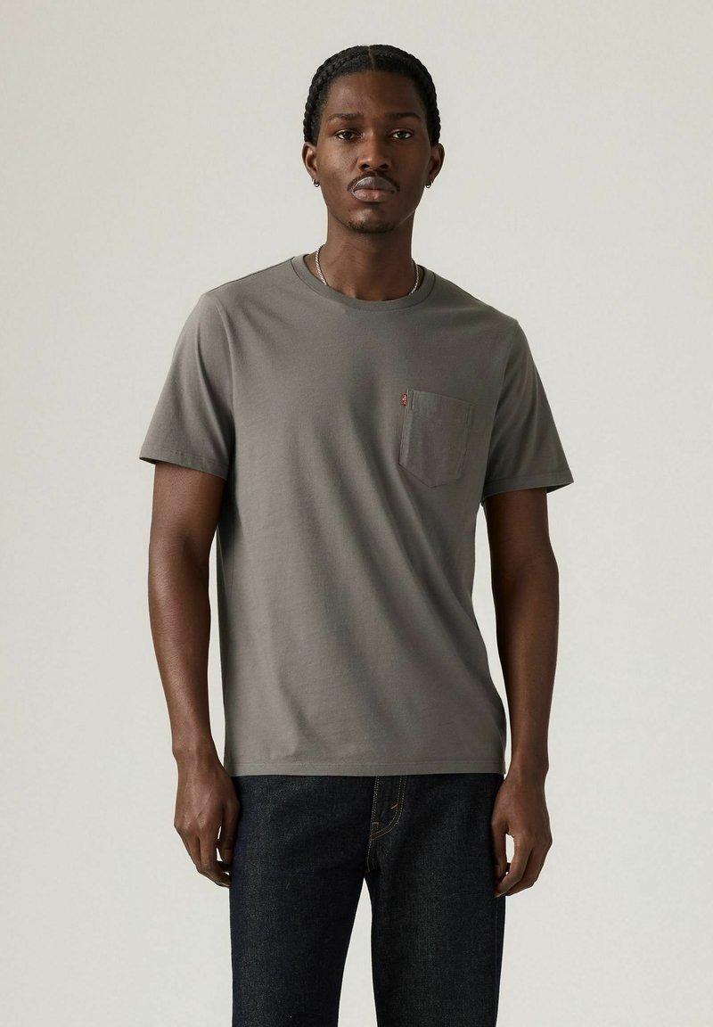 En ung mand står midt i billedet iført en grå kortærmet t-shirt med en brystlomme, parret med mørke jeans og en neutral baggrund.