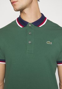 Zelená polo košile s texturovaným povrchem, která má námořnický límec s červenými a bílými pruhy a tři knoflíky. Logo Lacoste na hrudi.