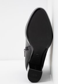 Semelle d'une bottine en cuir noir avec talon bloc, embout de talon en caoutchouc antidérapant et fermeture éclair latérale sur fond blanc.