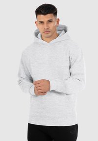 Grauer Kapuzenpullover aus weichem Stoff. Verfügt über eine Taschenfront, gerippte Bündchen und Saum. Das Model posiert mit verschränkten Armen.