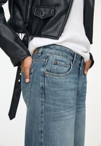 Blauwe spijkerbroeken van denim met een vervaagde afwerking, vijfzakkenontwerp, koperen klinknagels en een leren label op de achterkant. Gedragen met een wit t-shirt en een zwarte jas.