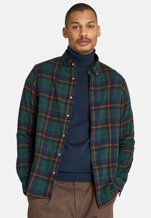 Camisa de cuadros verdes en tela de franela, con líneas naranjas, cuello con botones y botones plateados. Lleva puesta sobre un cuello de tortuga azul marino.