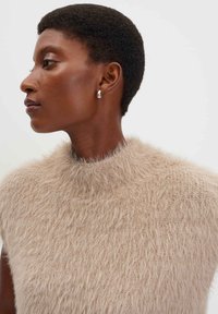 Maglione senza maniche beige realizzato in morbido e soffice tessuto a maglia; presenta un colletto alto e testurizzato e un design minimalista. Orecchino in argento mostrato.