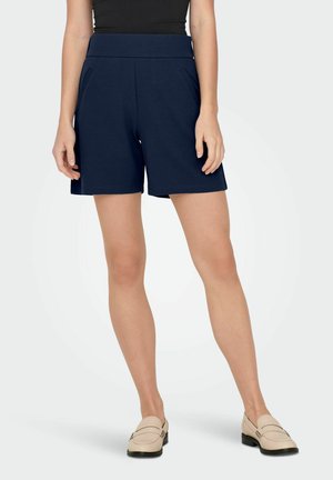 Donna che indossa pantaloni corti a vita alta blu navy con tasche laterali, abbinati a mocassini beige e una maglietta nera infilata, in piedi su uno sfondo bianco.