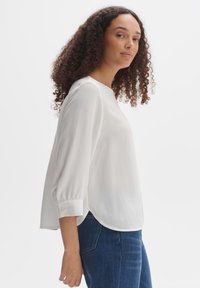 Blouse blanche avec un ourlet arrondi et des manches trois-quarts. Caractérisée par un tissu texturé et des poignets boutonnés. Portée avec un jean bleu.