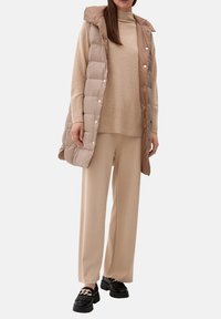 Femme portant un pull en maille beige, un pantalon à jambes larges, une veste matelassée sans manches avec capuche de couleur fauve, et des mocassins noirs ornés d'une chaîne métallique.