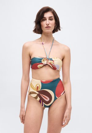 Kobieta w wielokolorowym, abstrakcyjnym, bezramkowym bikini z wysokim stanem, stojąca na tle gładkiego, jasnego tła.