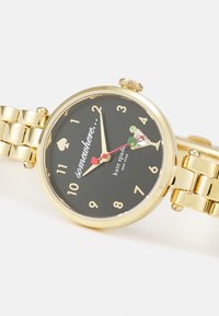 Gold-tone wristwatch with a black dial, featuring gold numbers and a heart symbol. Decorative cocktail glass design includes colored accents.