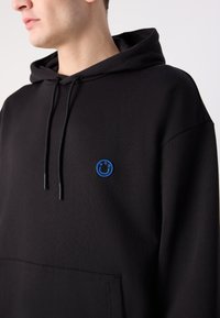 Jeune personne portant un sweat à capuche noir avec des cordons et un petit logo de visage souriant bleu sur la partie gauche de la poitrine.