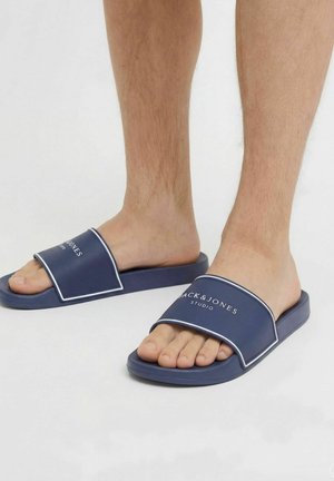 Blote voeten met marineblauwe Jack & Jones Studio slides op een witte vloer.