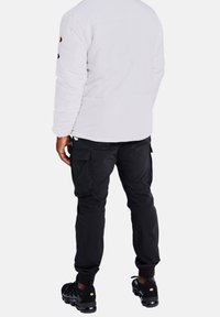 Person som bär en vit vadderad jacka, svarta cargobyxor och svarta sneakers står med avslappnade händer vid sidorna, vänd bort.