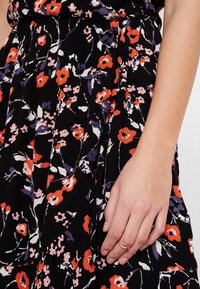 Robe noire à fleurs avec des fleurs rouges et violettes, tissu léger, détail de taille froncée et texture douce. Main avec une bague en or visible.