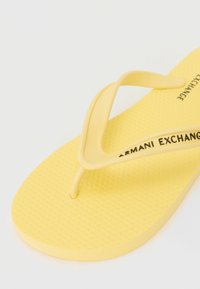 Gul gummiflip-flop med teksturert såle, med en tynn stropp og "ARMANI EXCHANGE" trykt på oversiden.