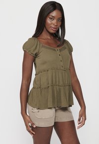 Blusa verde oliva con maniche corte a sbuffo, chiusura con bottoni e design a balze a strati. Abbinata a pantaloni corti beige chiaro con tasche.