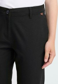 Black pants with a smooth texture, featuring a button closure and a small brown logo tab near the waistband. Tailored, slim fit design.