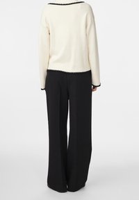 Cremfarbener Pullover mit schwarzem Wellensaum, kombiniert mit lockeren schwarzen Hosen. Der Stoff erscheint weich und strukturiert, mit einem klaren Design.