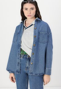 Denim srajca z jakno v srednje modri barvi, z gumbnim zapiranjem spredaj, dvema žepoma na prsih in sproščenim krojem. Oblečena čez svetlo sivo polo majico z črtami.