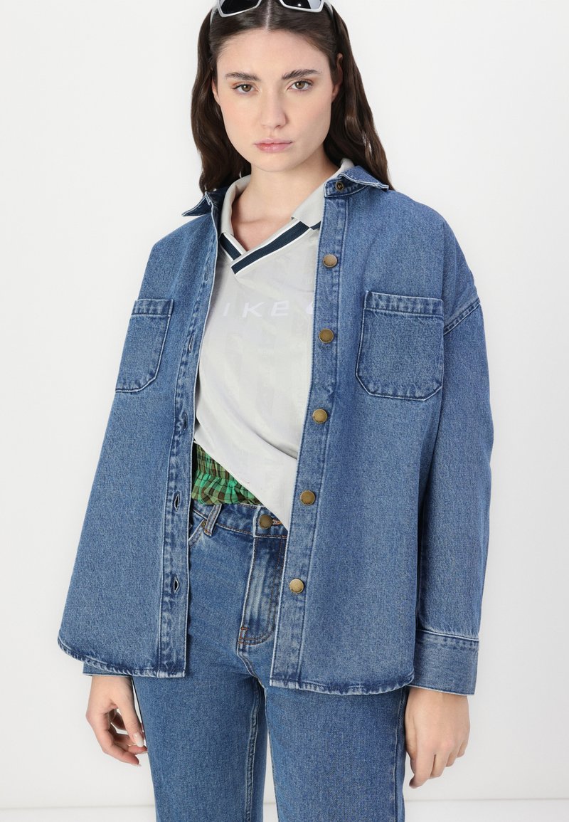 Camisa chaqueta de mezclilla en azul medio, con cierre de botones, dos bolsillos en el pecho y un corte relajado. Llevada sobre un polo gris claro a rayas.