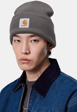 Gorro de punto acanalado gris con un parche del logo de Carhartt. Presenta un ajuste ceñido, textura suave y un diseño casual, llevado con una chaqueta de mezclilla.