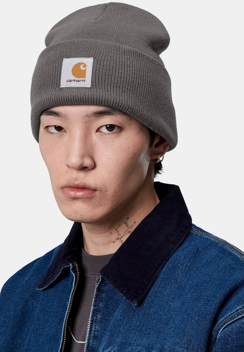 Gorro de punto acanalado gris con un parche del logo de Carhartt. Presenta un ajuste ceñido, textura suave y un diseño casual, llevado con una chaqueta de mezclilla.