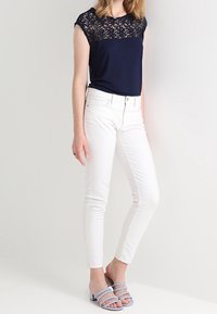 Kvinna i marinblå spetstopp, vita slim-fit byxor och ljusblå öppetåiga klacksandaler, står framför en enfärgad bakgrund.