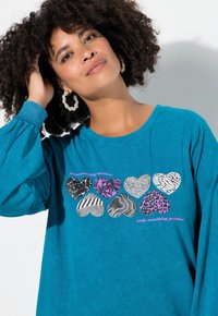 Kobieta z kręconymi włosami, ubrana w turkusową koszulę z ośmioma wzorzystymi sercami i napisem "pomaluj coś bez słów, napisz coś bez sensu."