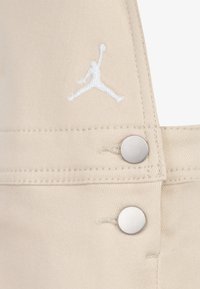 Bež kombinezon s izraženim bijelim vezom Jumpman logotipom, srebrnim metalnim gumbima i teksturiranim materijalom.